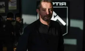 Mężczyzna o cynicznym spojrzeniu w czarnej koszuli, na tle logo z białym symbolem.