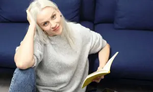 Kobieta z blond włosami, w szarym swetrze i jeansach, czyta książkę. To jak lalka streszczenie krótkie, relaksuje się na kanapie.