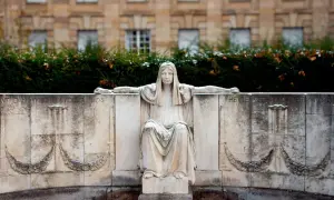 Kamienna figura siedzącej kobiety, symbolizująca fatum, z wyciągniętymi ramionami, na tle budynku i zieleni.