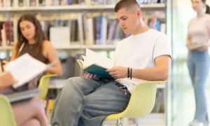Młody mężczyzna w białej koszulce pochłonięty lekturą w bibliotece. W tle inne osoby, półki z książkami. Czas jakby zatrzymał się w tym przedwiośniu.