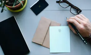 Dłoń trzyma ołówek, gotowa do napisania listu. Na stole leżą notes, koperta i papeteria z monogramem, czekające na adresata.