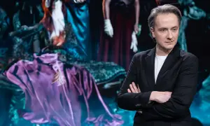 Mężczyzna w czarnym garniturze stoi na tle sceny z fioletową tkaniną i postaciami w strojach. Może to być nawiązanie do "Cichowskiego Dziadów".