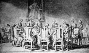Grupa ludzi przy stole, w strojach z epoki, z portretem na ścianie. Scena przypomina manieryzm w sztuce.