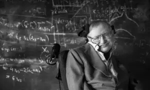 Młody Stephen Hawking w okularach, z uśmiechem, siedzi na wózku inwalidzkim przed tablicą pełną równań.