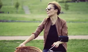 Kobieta w skórzanej kurtce i okularach przeciwsłonecznych na rowerze z wiklinowym koszykiem. Czasem warto zwolnić i docenić prostotę, zamiast ulegać konsumpcjonizmowi.