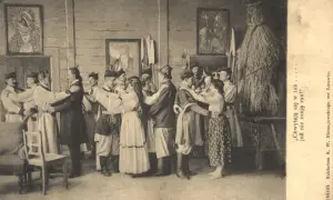 Grupa ludzi w strojach ludowych tańczy w pomieszczeniu. W tle obrazy i słomiana postać. To chocholi taniec.