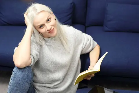 Kobieta z blond włosami, w szarym swetrze i jeansach, czyta książkę. To jak lalka streszczenie krótkie, relaksuje się na kanapie.