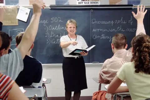 Nauczycielka w klasie, wskazując na tablicę, gdzie uczniowie podnoszą ręce. To praca u podstaw edukacji, budowanie przyszłości.