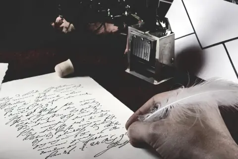 Dłoń z piórem pochyla się nad papierem z odręcznym pismem. Obok leży stempel, jakby czekał na zakończenie tej fraszki.