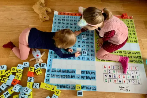 Dwie dziewczynki uczą się czytać, układając litery na planszy. Zabawa z literami wspiera naukę czytania.