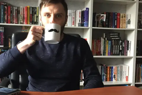 Adrian Bednarek z kubkiem w ręku, otoczony przez półki pełne książek.