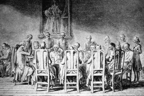 Grupa ludzi przy stole, w strojach z epoki, z portretem na ścianie. Scena przypomina manieryzm w sztuce.
