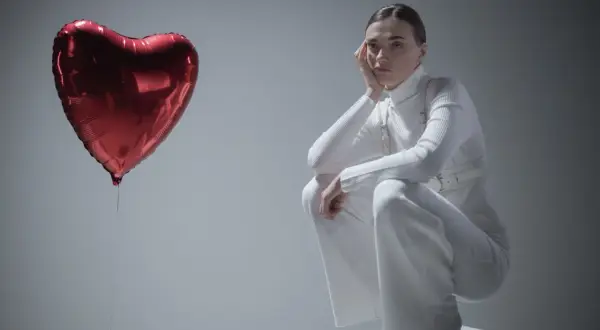 Kobieta w białym stroju siedzi na kostce, opierając głowę na dłoni. Obok niej unosi się czerwone serce-balon. Czy to symbol miłości, czy może zespół Otella?