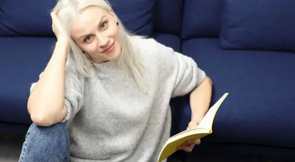 Kobieta z blond włosami, w szarym swetrze i jeansach, czyta książkę. To jak lalka streszczenie krótkie, relaksuje się na kanapie.