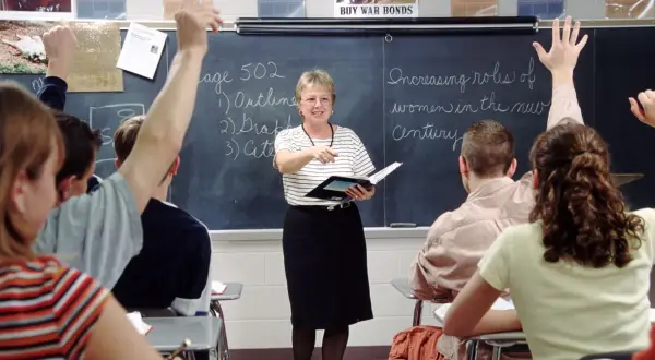 Nauczycielka w klasie, wskazując na tablicę, gdzie uczniowie podnoszą ręce. To praca u podstaw edukacji, budowanie przyszłości.