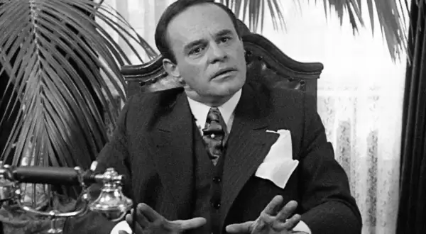 Nikodem Dyzma w eleganckim garniturze, z dłońmi uniesionymi w geście, siedzi przy biurku z zabytkowym telefonem.