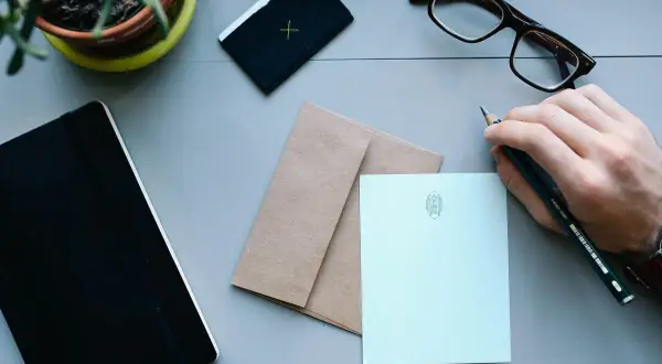 Dłoń trzyma ołówek, gotowa do napisania listu. Na stole leżą notes, koperta i papeteria z monogramem, czekające na adresata.