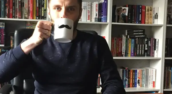 Adrian Bednarek z kubkiem w ręku, otoczony przez półki pełne książek.