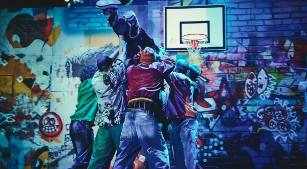Tancerze w stylu hip-hop unoszą kolegę na tle ściany z graffiti, przypominającej motywy z Ferdydurke.