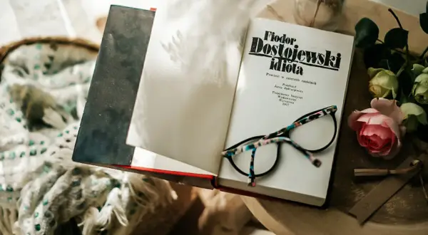 Okładka książki "Fiodor Dostojewski idiota" leży otwarta obok okularów i róży.