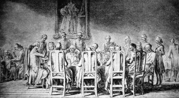 Grupa ludzi przy stole, w strojach z epoki, z portretem na ścianie. Scena przypomina manieryzm w sztuce.