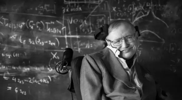 Młody Stephen Hawking w okularach, z uśmiechem, siedzi na wózku inwalidzkim przed tablicą pełną równań.