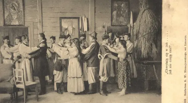 Grupa ludzi w strojach ludowych tańczy w pomieszczeniu. W tle obrazy i słomiana postać. To chocholi taniec.