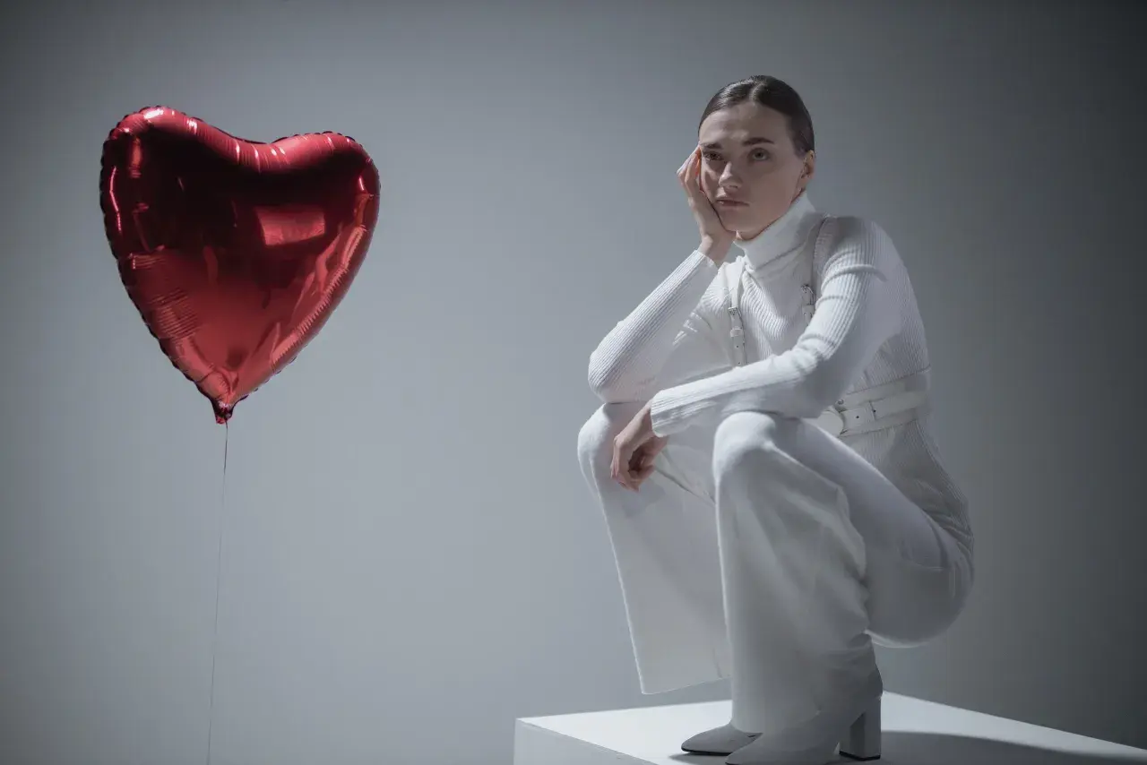 Kobieta w białym stroju siedzi na kostce, opierając głowę na dłoni. Obok niej unosi się czerwone serce-balon. Czy to symbol miłości, czy może zespół Otella?