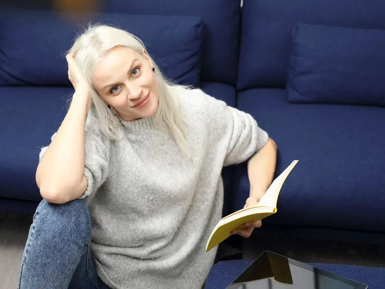 Kobieta z blond włosami, w szarym swetrze i jeansach, czyta książkę. To jak lalka streszczenie krótkie, relaksuje się na kanapie.