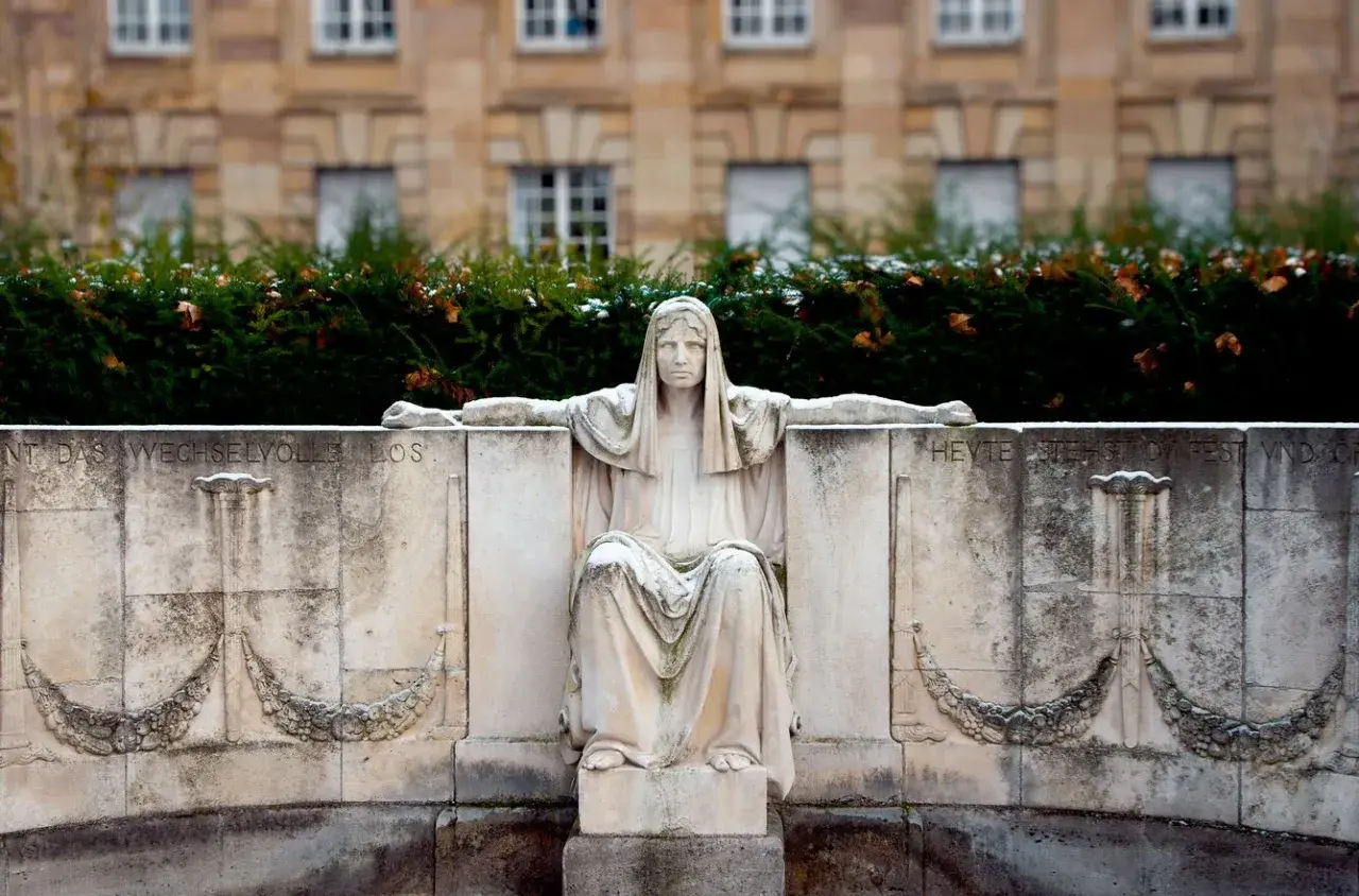Kamienna figura siedzącej kobiety, symbolizująca fatum, z wyciągniętymi ramionami, na tle budynku i zieleni.