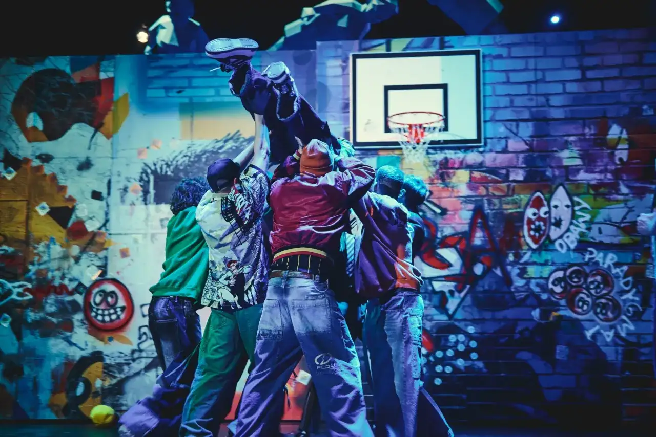 Tancerze w stylu hip-hop unoszą kolegę na tle ściany z graffiti, przypominającej motywy z Ferdydurke.