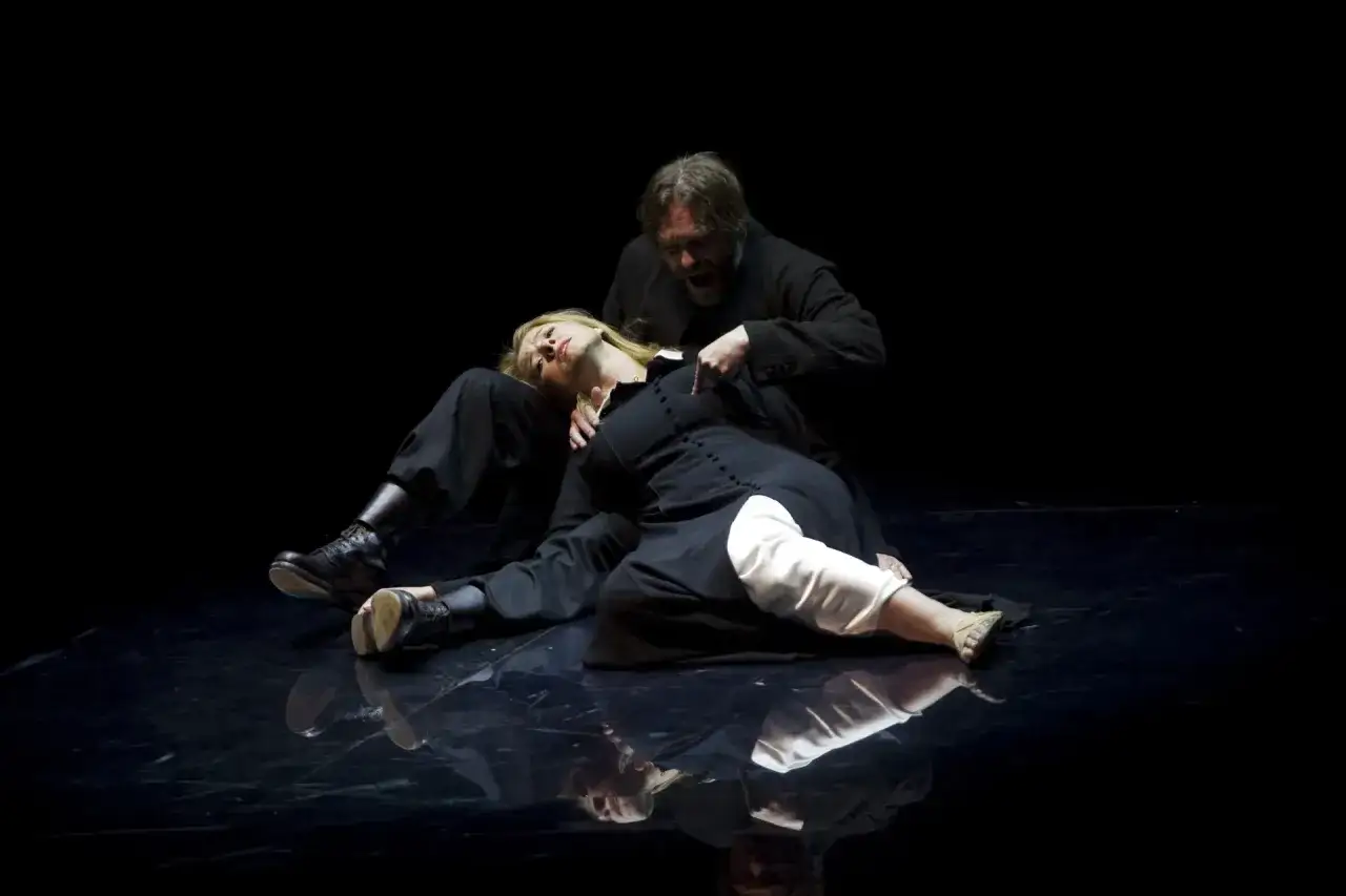Mężczyzna w dramatycznym geście wskazuje na kobietę leżącą na scenie. Scena z przedstawienia