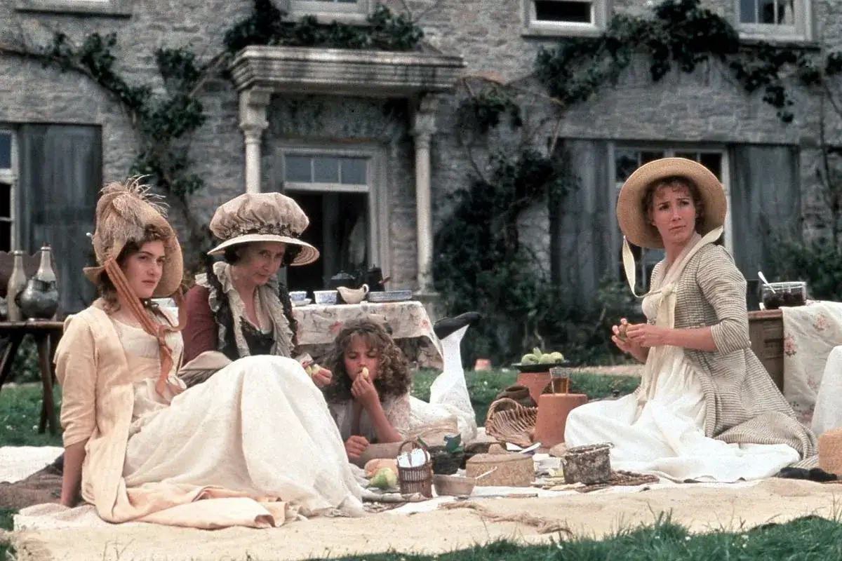 Kobiety w strojach z epoki, przypominające rodzeństwo Jane Austen, piknikują przed kamiennym domem.