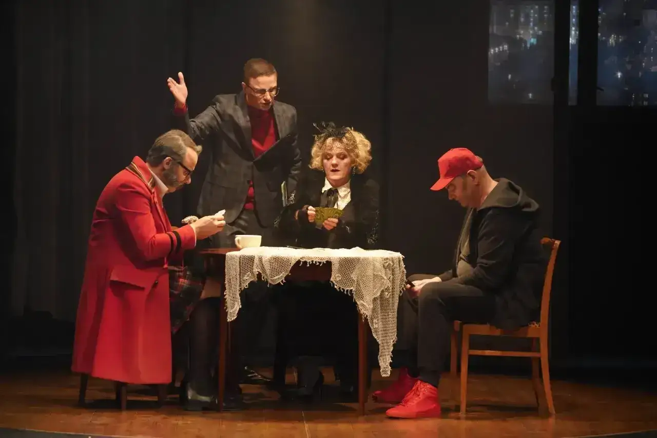 Cztery postacie, w tym kobieta w czarnej sukni i kapeluszu, siedzą przy stole. Grają w karty, a mężczyzna w czerwonym płaszczu czyta list.