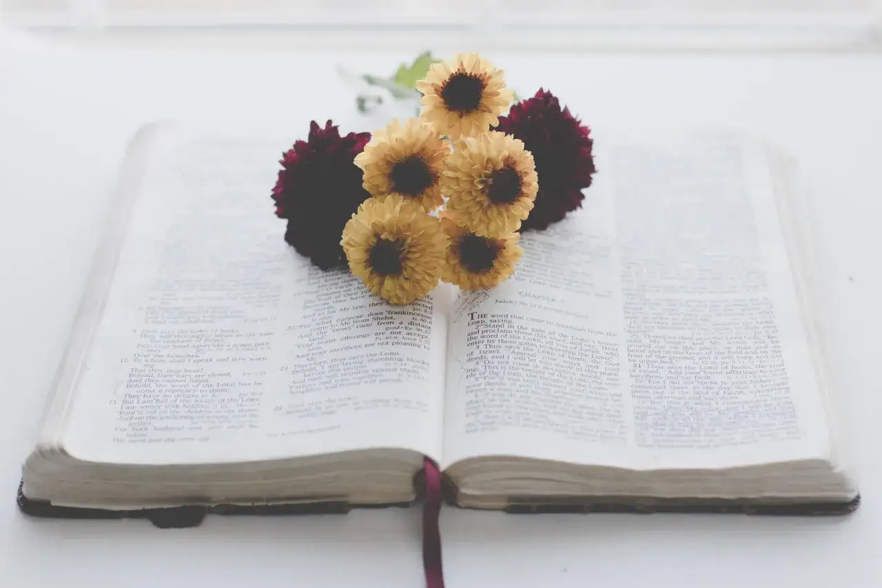 Otwarta Biblia z kwiatami na stronach. Tekst przypomina epopeję, opowiadając historie o wierze i ludzkich losach.