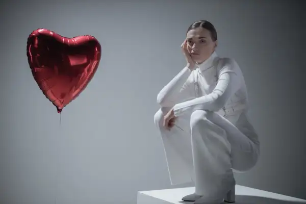 Kobieta w białym stroju siedzi na kostce, opierając głowę na dłoni. Obok niej unosi się czerwone serce-balon. Czy to symbol miłości, czy może zespół Otella?