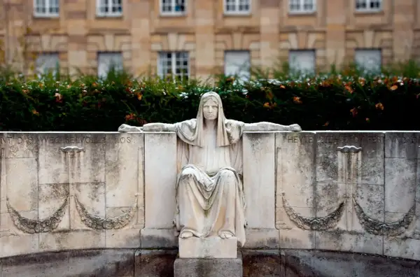 Kamienna figura siedzącej kobiety, symbolizująca fatum, z wyciągniętymi ramionami, na tle budynku i zieleni.